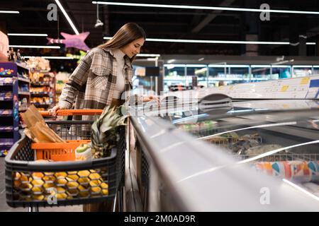 Eine junge Frau mit Einkaufswagen sucht sich das Essen im Tiefkühlbereich eines Supermarkts aus Stockfoto