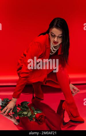 Elegante Frau in Perlenkette, die neben Rose auf reflektierender Oberfläche auf rotem Hintergrund posiert Stockfoto