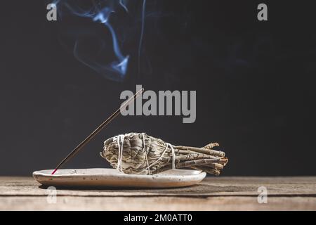 Brennender aromatischer Incense Smoky Stick und ein Bündel Salvia Salvia Salbe auf dem Keramikhalter für Energiereinigung und Entspannungstherapie. Rustikales Holz und dunkles B Stockfoto