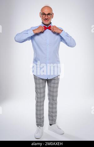 Voller glücklicher Mann, der auf isoliertem weißen Hintergrund steht und seine Fliege justiert. Lächelnder weißer Mann mit Fliege und karierter Hose. Co Stockfoto