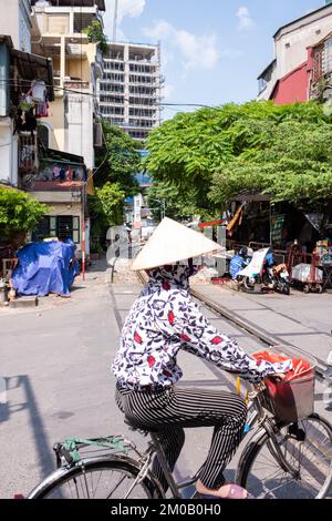 Hanoi, Vietnam - 16. September 2018: Hübsche vietnamesische Frau mit traditionellem Kopfschmuck überquert die Bahnstrecke auf einem Fahrrad Stockfoto