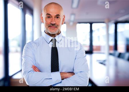 Portrait eines selbstbewussten, männlichen CEO, der im Büro steht. Ein erfahrener Manager trägt Hemd und Krawatte. Ein Geschäftsmann, der mit gefalteten Armen steht und guckt Stockfoto