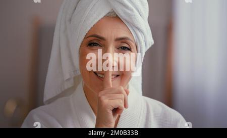 Alte, weiße, glückliche Frau, ältere Dame, weibliche Model mit Handtuch auf dem Kopf im Bademantel, die auf ein Lächeln der Kamera schaut und eine Geste der Stille zeigt Stockfoto