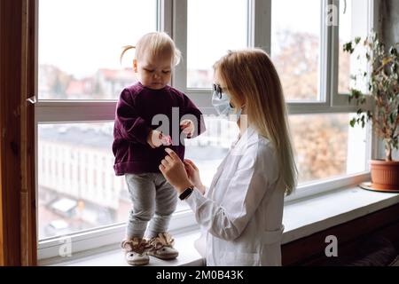 Kleines blondes Mädchen, das auf dem Fensterbrett steht, das Spielzeug in der Hand in der Nähe von Kinderärzten sieht, die zu Hause weiße Uniform tragen. Stockfoto