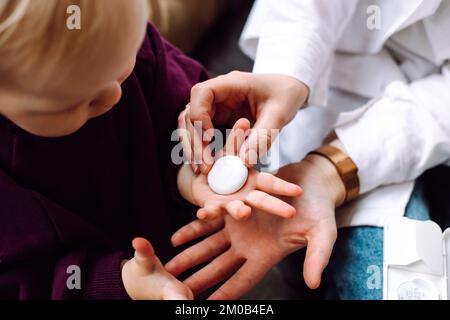 Draufsicht eines erstaunlichen kleinen Kindes, das auf dem Sofa sitzt und die Handfläche für ein weißes Thermometer zur Temperaturmessung öffnet. Stockfoto