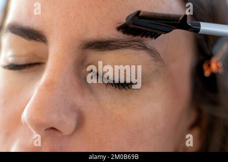 Nahaufnahme eines jungen Weibchens mit geschlossenen Augen, das die Augenbrauen mit dem Kamm putzt, während es morgens Make-up macht Stockfoto