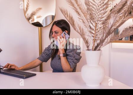Junge Frau, Rezeptionistin eines Schönheitssalons, geht ans Telefon, während sie auf der Computertastatur tippt Stockfoto