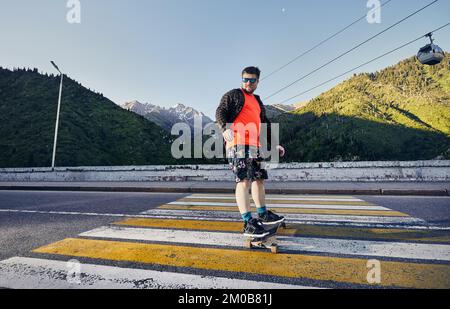 Bärtige Skater fahren auf der Bergstraße auf seinem Longboard in Medeo Dum Almaty, Kasachstan Stockfoto