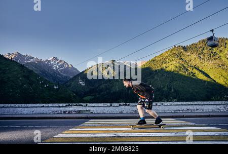 Bärtige Skater fahren auf der Bergstraße auf seinem Longboard in Medeo Dum Almaty, Kasachstan Stockfoto