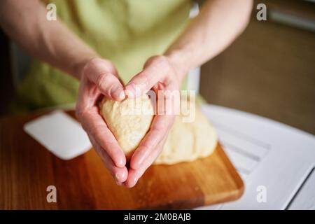 Arbeiten zu Hause Küchenkonzept, hausgemachtes Backen. Der Küchenchef in grüner Schürze hält die Teigkugeln in Händen, die in Herzform gefaltet sind. Hochwertiges Foto Stockfoto