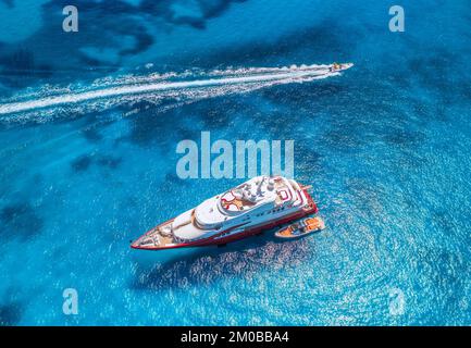 Blick aus der Vogelperspektive auf die wunderschöne rote Luxusyacht und das Boot im blauen Meer bei Sonnenuntergang im Sommer. Insel Sardinien, Italien. Blick von oben auf Schnellboot, Küste, Transpa Stockfoto