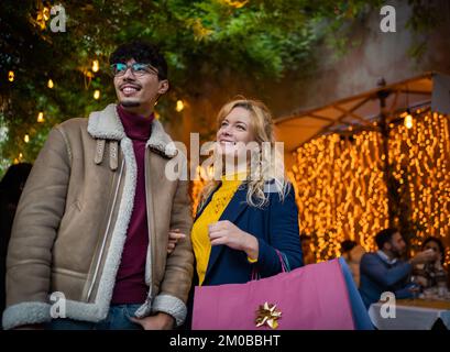 Ein junges, multiethnisches Paar, das lächelt und die Leute ansieht, während es Einkaufstüten in der Hand hält Stockfoto