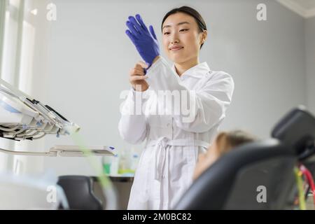 Wunderschöne asiatische Zahnärztin trägt blaue Gummihandschuhe, steht in einer Zahnarztpraxis, bereit für die Untersuchung und Behandlung ihrer Patientin. Glücklicher Patient und Zahnarztkonzept Stockfoto