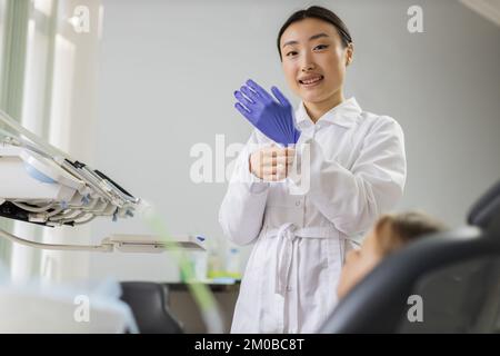 Wunderschöne asiatische Zahnärztin trägt blaue Gummihandschuhe, steht in einer Zahnarztpraxis, bereit für die Untersuchung und Behandlung ihrer Patientin. Glücklicher Patient und Zahnarztkonzept Stockfoto