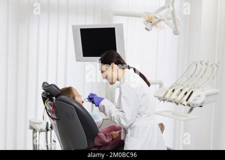 Schönes Lächeln mit weißen Zähnen. Der asiatische Zahnarzt untersucht die Mundhöhle eines jungen schönen Mädchens durch eine Lupe in der Zahnarztpraxis. Stockfoto