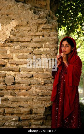 Balkh, Provinz Balkh/Afghanistan: Ein afghanisches Mädchen in einem glitzernden roten Kleid steht an einer alten Backsteinmauer in Balkh, Nordafghanistan. Stockfoto