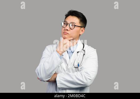 Nachdenklicher, reifer chinesischer Therapeut in weißem Mantel und Brille denkt und schaut sich freien Raum an Stockfoto