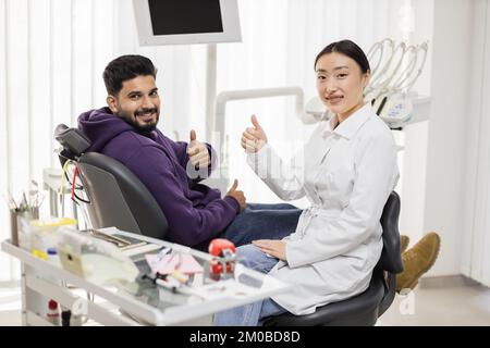 Zahnbehandlung im Zahnkrankenhaus. Lächelnder bärtiger Mann, der auf dem Zahnarztstuhl in einer hellen modernen Zahnklinik sitzt, der aufgeregten schönen asiatischen Zahnärztin Daumen hoch zeigt und in die Kamera schaut. Stockfoto