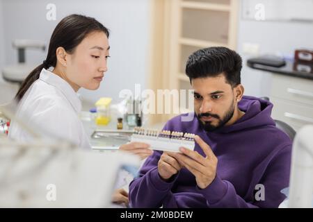 Zahnzähntonpalette. Wunderschöne asiatische weibliche Stomatologin Kieferorthopädie Zahnärztin wählt Farbe des Patienten attraktive bärtige männliche Zähne für die Aufhellung in der Klinik. Stockfoto