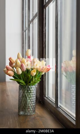 Wunderschöner Strauß bunter Tulpen in Glasvase auf Holzfenstern in der Nähe des Fensters Stockfoto