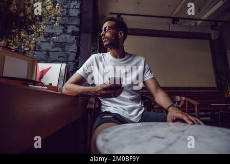 Ein afroamerikaner in einem weißen T-Shirt sitzt in einem Café und telefoniert. Modell. Stockfoto