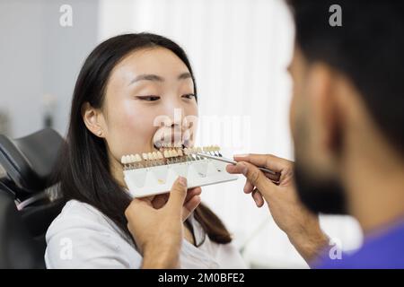 Zahnarzt wählt Füllungsfarbe aus Palette. Rückblick des männlichen Zahnarztes bei der Prüfung und Farbwahl junger asiatischer Frauenzähne im Innenbereich des modernen Krankenhauses. Stockfoto