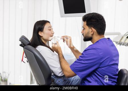 Zahnarzt wählt Füllungsfarbe aus Palette. Rückblick des männlichen Zahnarztes bei der Prüfung und Farbwahl junger asiatischer Frauenzähne im Innenbereich des modernen Krankenhauses. Stockfoto