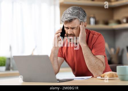 Gestresster Geschäftsmann mittleren Alters, der von zu Hause aus arbeitet, telefoniert Stockfoto