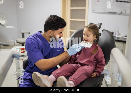 Zahnzähntonpalette. Stomatologe Kieferorthopäde männlicher Zahnarzt wählt die Farbe der süßen Zähne des kleinen Vorschulmädchens für die Aufhellung in der Klinik aus. Stockfoto