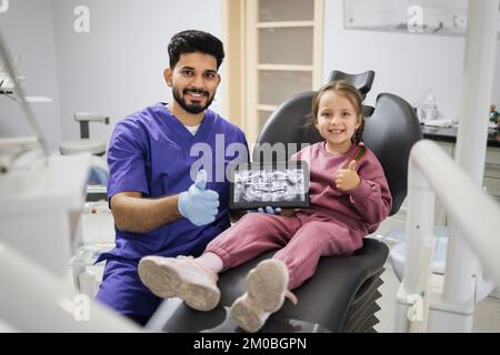 Hübsches lächelndes Vorschulmädchen, sitzt auf dem Zahnarztstuhl und sieht sich ein digitales Tablet an, mit einem Röntgenbild der Zähne, zusammen mit ihrem professionellen bärtigen Zahnarzt in der Klinik, der Daumen nach oben zeigt. Stockfoto