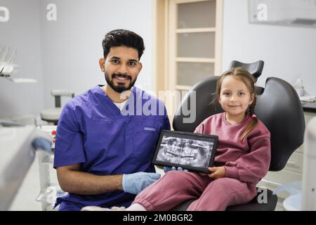 Hübsches, lächelndes, weißes Vorschulmädchen, sitzt auf einem Zahnarztstuhl und sieht sich ein digitales Tablet an, mit einem Röntgenbild der Zähne, zusammen mit ihrem professionellen, bärtigen Zahnarzt in der Klinik. Stockfoto