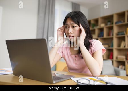 Traurige, müde asiatische Dame mit Laptop, die Kopfschmerzen hatte, die Hände an Schläfen drückte, am Schreibtisch im Wohnzimmer saß Stockfoto