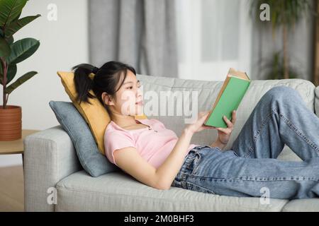 Sorgenfreie koreanische Dame, die auf der Couch im Wohnzimmer liegt, Buch liest, während sie sich am Wochenende zu Hause ausruht, Kopierraum Stockfoto