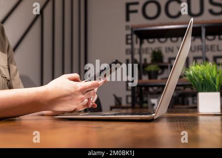 Smartphone in der Hand, Sitz am hölzernen Büroschreibtisch mit Laptop, PC. Arbeiten mit Informationstechnologien, Geräten. Hochwertiges Foto Stockfoto