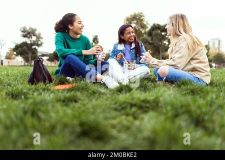 Drei junge Schülerinnen essen zu Mittag und sitzen auf dem Rasen am Campus College Stockfoto