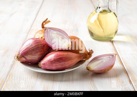 Ganze und halbierte rote Schalotten auf einem Teller über einem weißen Holztisch. Ungeschälte Eflöten-Zwiebeln aus der Nahaufnahme. Frische, rohe, lange Zwiebeln, bereit zum Kochen. Vorderansicht Stockfoto