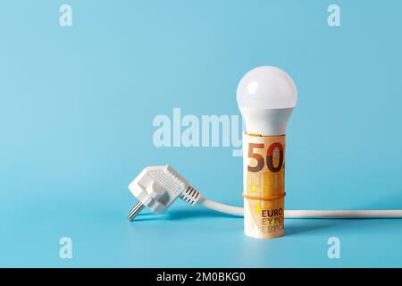 LED-Glühbirne auf gerollten 50-Euro-Banknoten und weißer Netzstecker auf blauem Hintergrund. Steigende Energiekosten, Energiekrise und Inflationskonzept. Stockfoto