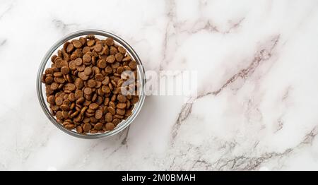 Trockenes Haustierfutter in einer Glasschüssel mit Marmorhintergrund. Glasfütterungsschüssel mit glutenfreien, trockenen Eiweißkibbles für die Nahaufnahme von Katzen. Vollständiges Haustierfutter. Stockfoto