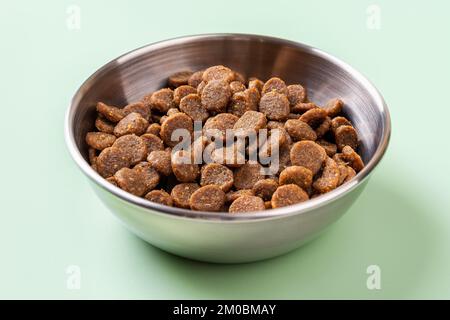 Trockenes Tierfutter in einer Metallschüssel auf grünem Hintergrund. Glutenfreie trockene Eiweißkibbles für Katzen in einer Edelstahl-Fütterschüssel. Vorderansicht. Stockfoto