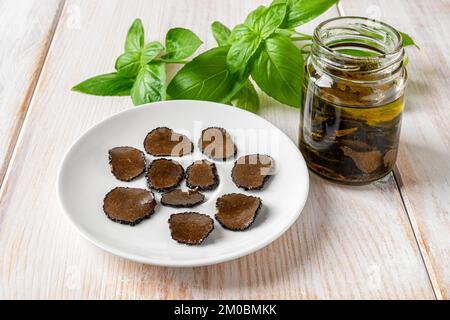 Trüffeln Sie Carpaccio-Scheiben auf einem Teller und einem Glas mit in Öl geschnittenem schwarzem Trüffel auf einem weißen Holztisch. Dosenpilze. Gourmet-Geschmack Stockfoto