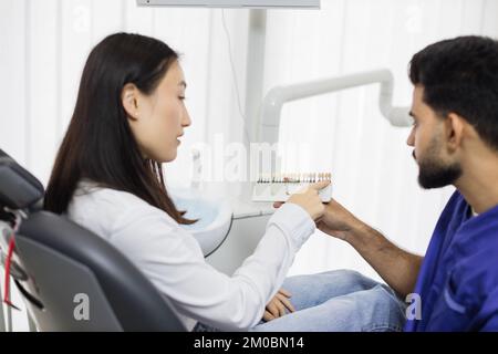 Seitennahansicht eines attraktiven männlichen Zahnarztes zur Prüfung und Farbwahl junger asiatischer Frauenzähne im Innenbereich in einer modernen, leichten Krankenhauspraxis. Stockfoto