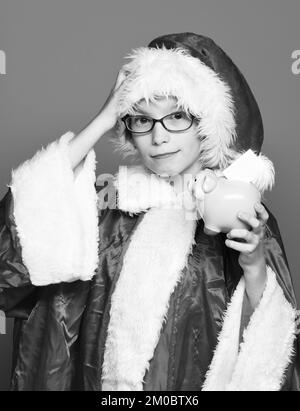 Junger, süßer weihnachtsmann mit Brille in rotem Pullover und Neujahrsmütze mit rosa Schweinebank mit erhobener Hand auf blauem Studio Stockfoto