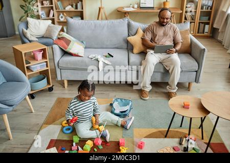 Großer Blickwinkel auf einen schwarzen Jungen, der mit Spielzeugen auf dem Boden spielt, während der Vater im Hintergrund arbeitet, Kopierbereich Stockfoto