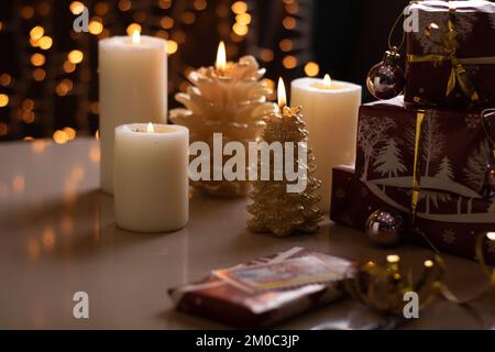 Mit Fotos verpackte Schachteln mit Geschenken stehen auf einem Tisch mit beleuchteten Kerzen im Hintergrund, mit festlicher Girlande Stockfoto