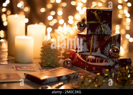 Fotogeschenke stehen in einer vertikalen Reihe, in festliches Papier eingewickelt, auf dem Tisch neben brennenden Kerzen, im Hintergrund ein Bokeh aus festlichem Weihnachtsfest Stockfoto