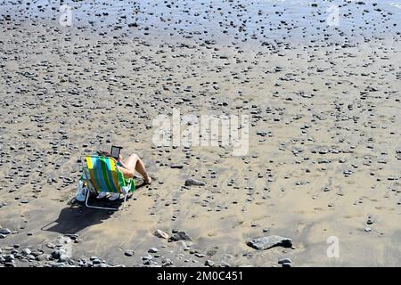Sonnenbaden am Sandstrand mit Kieselsteinen, sitzen auf einem Klappstuhl und lesen mit einem Hauch von Meer, Playa del Águila, Gran Canaria Stockfoto