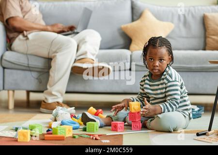 Gesamtporträt eines süßen schwarzen Kleinkindes, das mit Spielzeugblöcken spielt, während er zu Hause auf dem Boden sitzt und der Vater im Hintergrund arbeitet, Kopierbereich Stockfoto