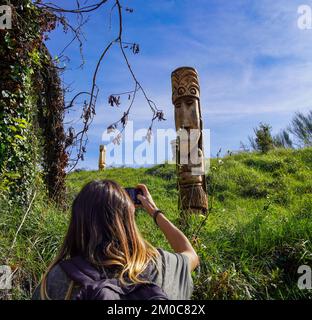 Langhaarige junge Frau, die ein Foto von einem hölzernen Moai macht, der im grünen Gras gebeugt ist. Die Skulptur befindet sich in einer erhöhten Position. Stockfoto