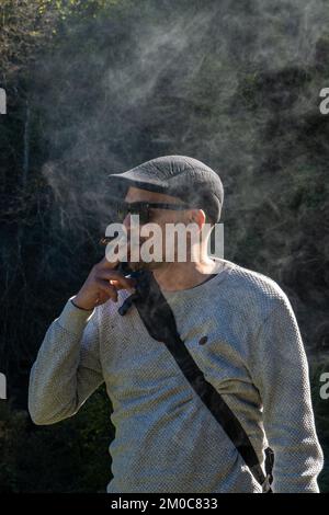 Moderner Mann und Künstler in einer Künstlermütze mit Sonnenbrille, die seinen Tabak raucht, erleuchtet von der untergehenden Sonne und umgeben von Rauch. Stockfoto