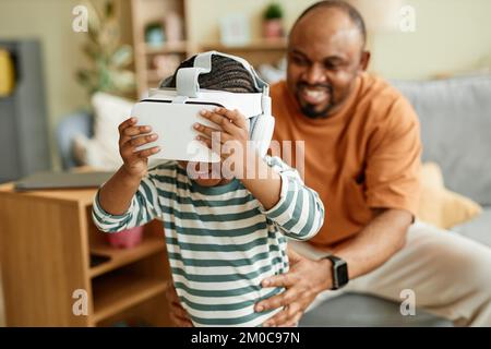 Porträt eines süßen schwarzen Kindes, das ein VR-Headset trägt und Virtual-Reality-Unterhaltung für Kinder mit Vater im Hintergrund genießt Stockfoto